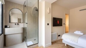 Ein modernes Hotelzimmer mit einer gläsernen Dusche, einem großen Spiegel, einem wandmontierten Fernseher und einem ordentlich bezogenen Bett.
