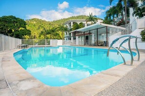 A swimming pool with a clear blue water, surrounded by a tiled deck, and a building with a covered patio area in the background. - Nalani On Airlie (Airlie Beach)
