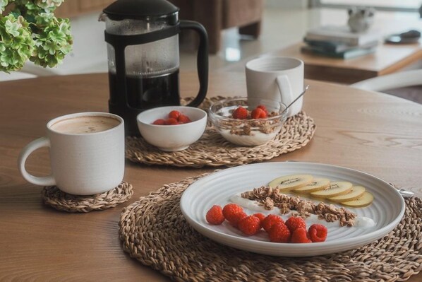Un desayuno con cafetera, dos tazas, un plato de frambuesas y fruta en rodajas, y un bol de granola.
