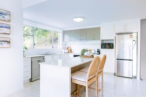 A modern kitchen with white cabinetry, a central island with chairs, a dishwasher, and a refrigerator. - Nalani On Airlie (Airlie Beach)