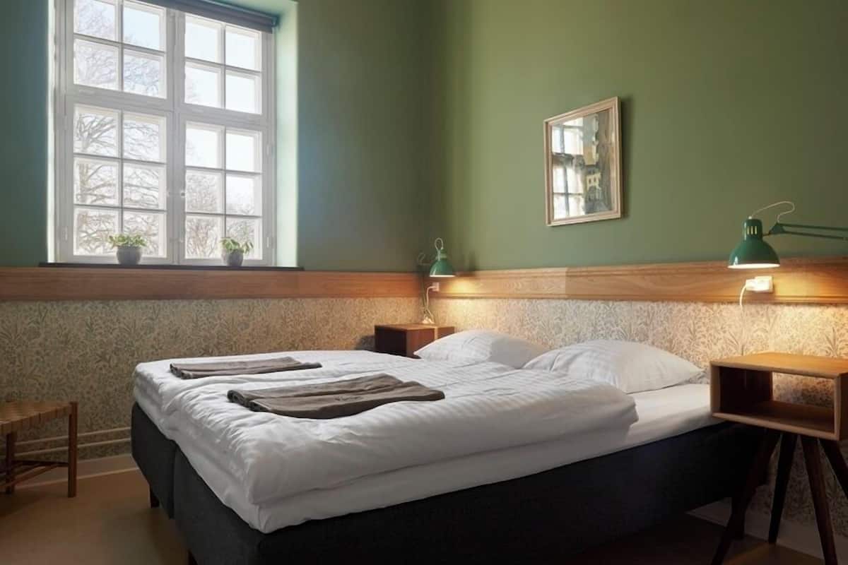 Un lit bien fait, avec des oreillers et une couverture, une table de chevet avec une lampe, une petite chaise et une fenêtre agrémentée de plantes en pot.