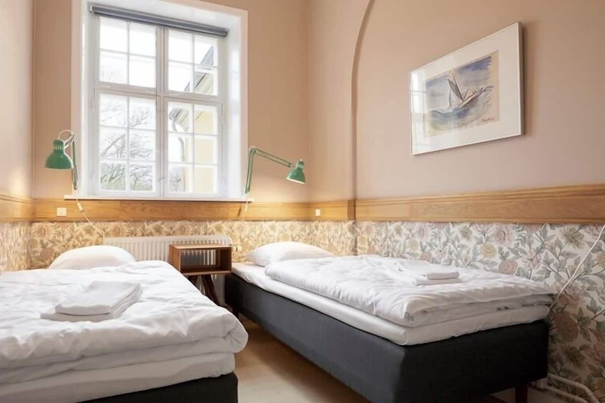 Une chambre d’hôtel avec deux lits, une fenêtre, un radiateur, un bureau et un tableau au mur.