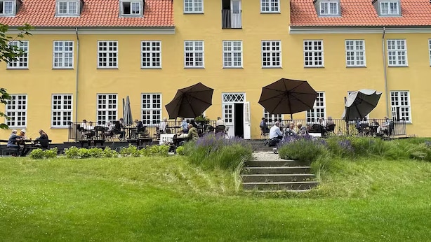Een geel gebouw met een rode pannendak, veel ramen en terras met parasols.