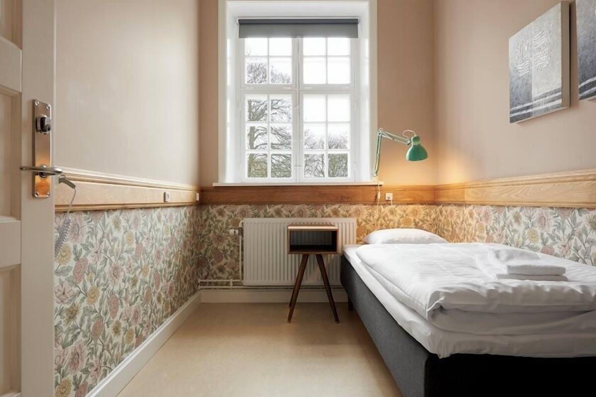 Une chambre à coucher avec un lit, un radiateur, une fenêtre avec des stores et une petite table.