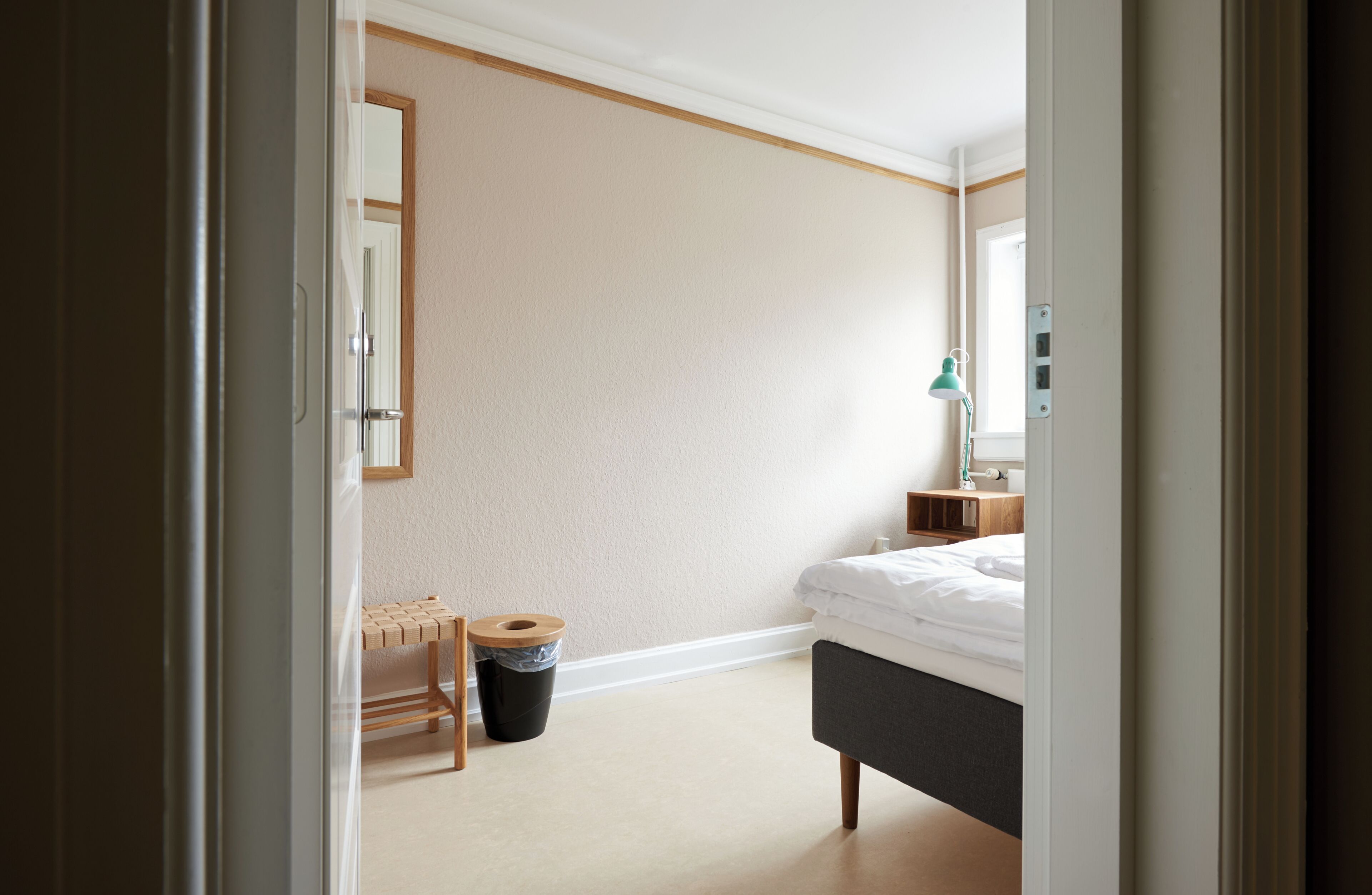 Un dormitorio con cama, mesita de noche, lámpara, cubo de basura y un banco de madera.