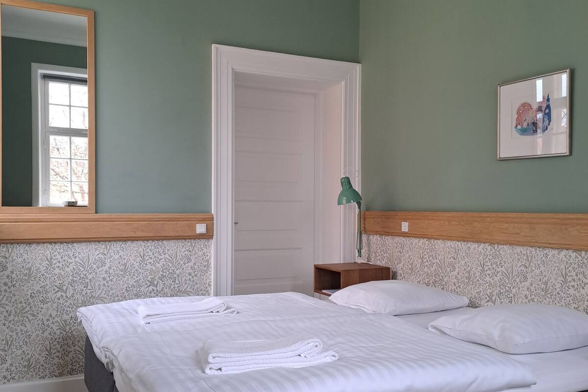 Un lit bien fait, avec du linge de lit et des serviettes blanches, une tête de lit en bois, une petite table de chevet avec une lampe, et une porte donnant sur une autre pièce.