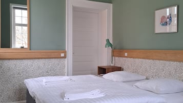A neatly made bed with white linens and towels, a wooden headboard, a small bedside table with a lamp, and a door leading to another room.