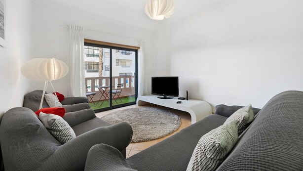Un soggiorno moderno con divani grigi, una TV a schermo piatto, una lampada da terra e vista su un balcone con mobili da esterno.