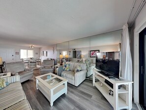 A living room with a sofa, armchairs, a coffee table, a TV, and a large mirror. - Bent Palm 403 | Poolside + Beach Views (Ormond Beach)
