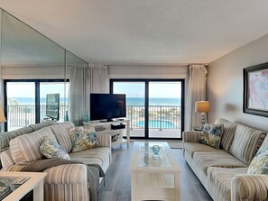 A hotel room with a large window, a TV, a sofa, a coffee table, and a lamp. - Bent Palm 403 | Poolside + Beach Views (Ormond Beach)