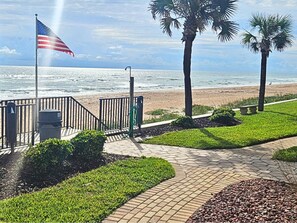 A paved walkway leading to a beach with palm trees and an American flag. - Bent Palm 403 | Poolside + Beach Views (Ormond Beach)