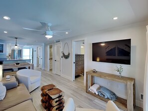 A modern living room with a sofa, wooden stools, a TV, and a fireplace. - Pointe South 106 | Newly Reno + Resort Pool Access (Fort Myers Beach)