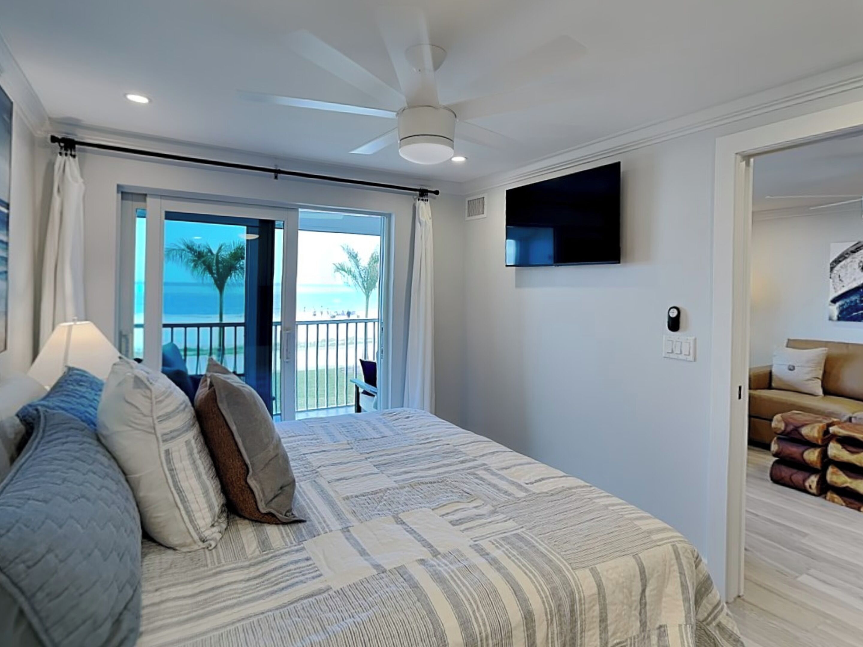 A bedroom with a bed, a ceiling fan, a TV, and a balcony with a view of the beach.