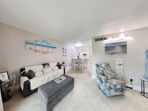 A living room with a sofa, a grey ottoman, and a patterned armchair. There is a dining area in the background with a table and chairs. - Bent Palm 221 | Pool and Beach Views (Ormond Beach)