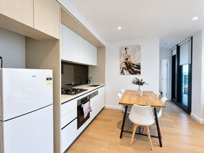 A modern kitchen with white cabinetry, a built-in oven, and a dining area with a wooden table and white chairs. - Luxury 2BR2BA Apt MQ+ Pool+Gym in Mel  Docklands (Docklands)