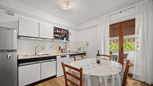 Eine Küche mit weißen Schränken, einem Esstisch mit Stühlen, einem Kühlschrank und einem Fenster mit Vorhängen.