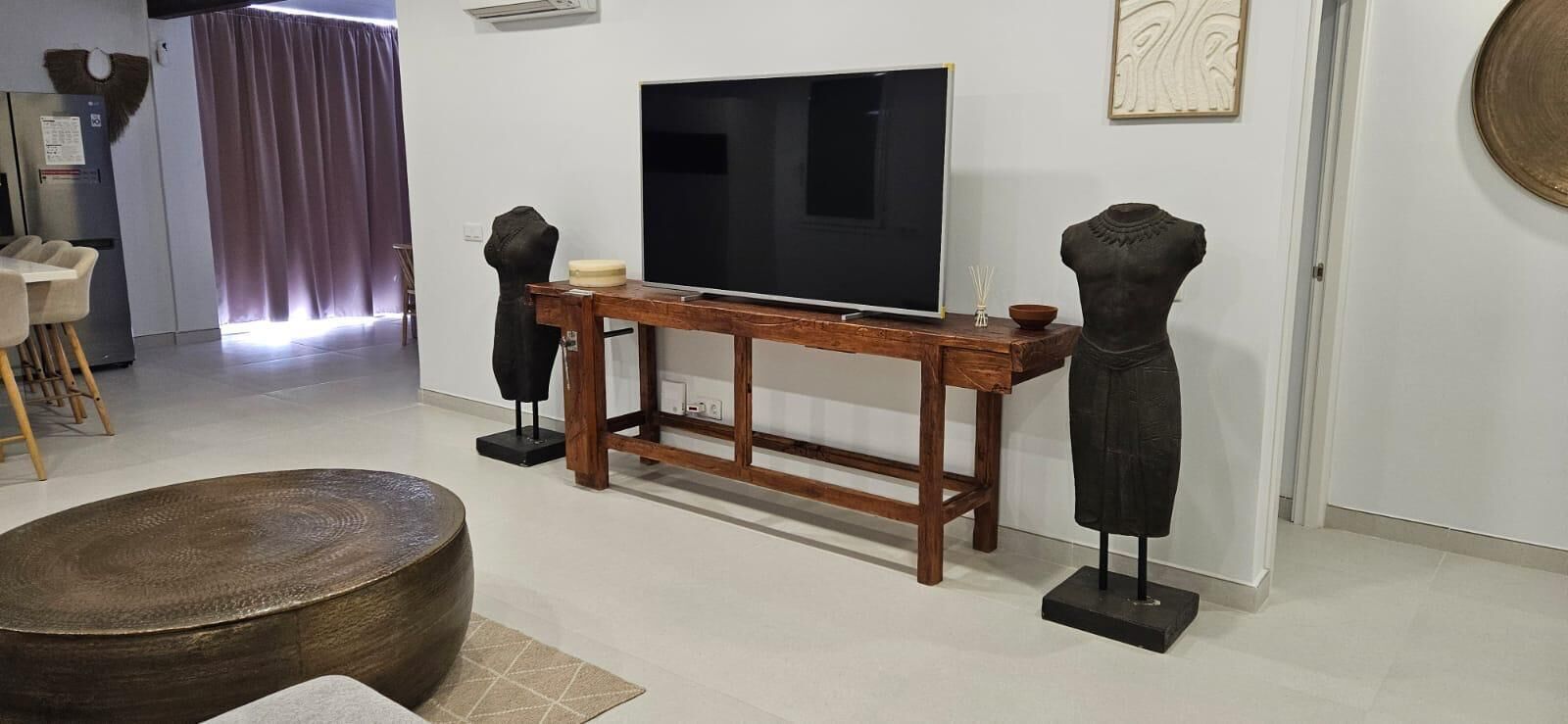 Ett vardagsrum med en tv på ett träställ, ett runt träbord och två svarta skulpturer på piedestaler.