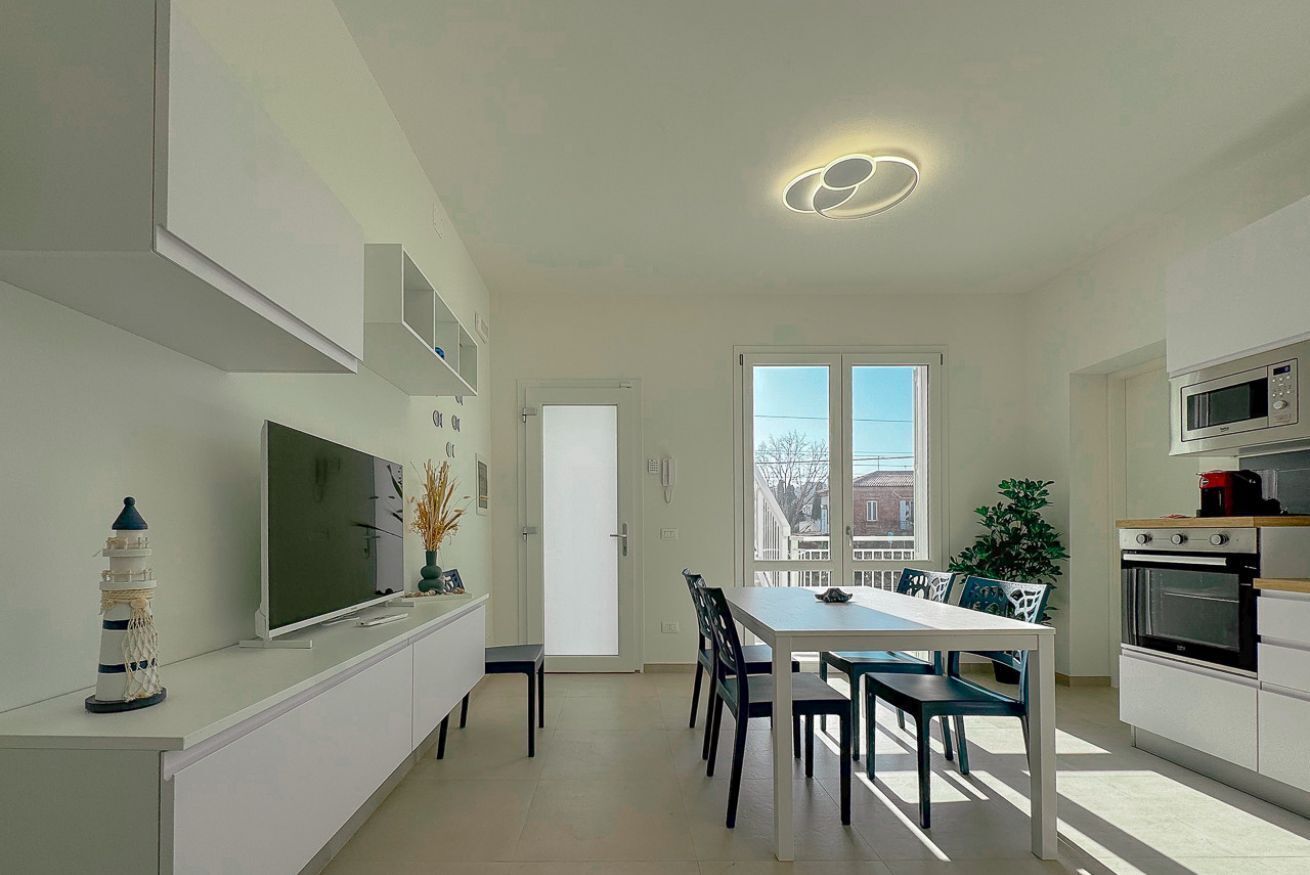 Una cocina moderna con armarios blancos, un comedor con mesa y sillas, un televisor sobre un mueble blanco y un amplio ventanal con vistas a edificios.