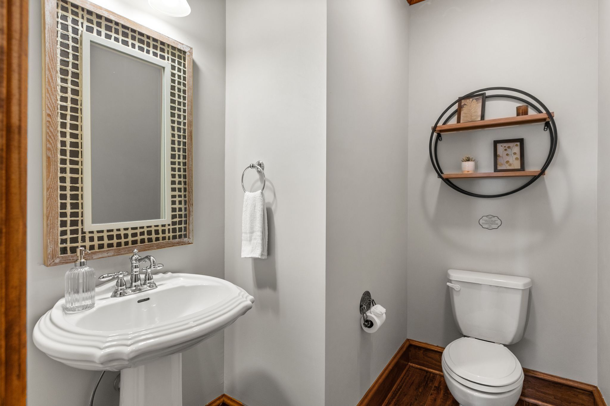 Un baño con un lavabo blanco, un espejo con marco de madera, un inodoro y una estantería con una imagen y una planta pequeña.