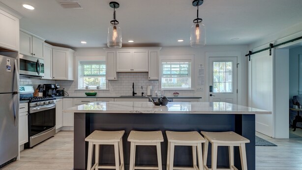 A modern kitchen with white cabinets, a large island with stools, stainless steel appliances, and pendant lighting. - A Perfect Place: Newly Remodeled Ground Level with Pool! (Holmes Beach)