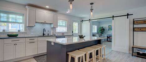 A modern kitchen with white cabinets, a large island with stools, pendant lighting, and a view into the adjacent living area.
