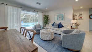 A living room with a wooden dining table, grey armchairs, a white sofa with blue cushions, a round ottoman, and a large window with curtains. - A Perfect Place: Newly Remodeled Ground Level with Pool! (Holmes Beach)