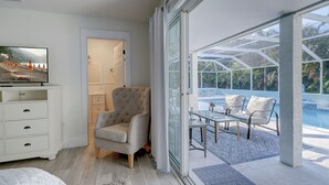 A modern hotel room with a TV, a comfortable armchair, a glass door leading to a pool area with outdoor seating. - A Perfect Place: Newly Remodeled Ground Level with Pool! (Holmes Beach)
