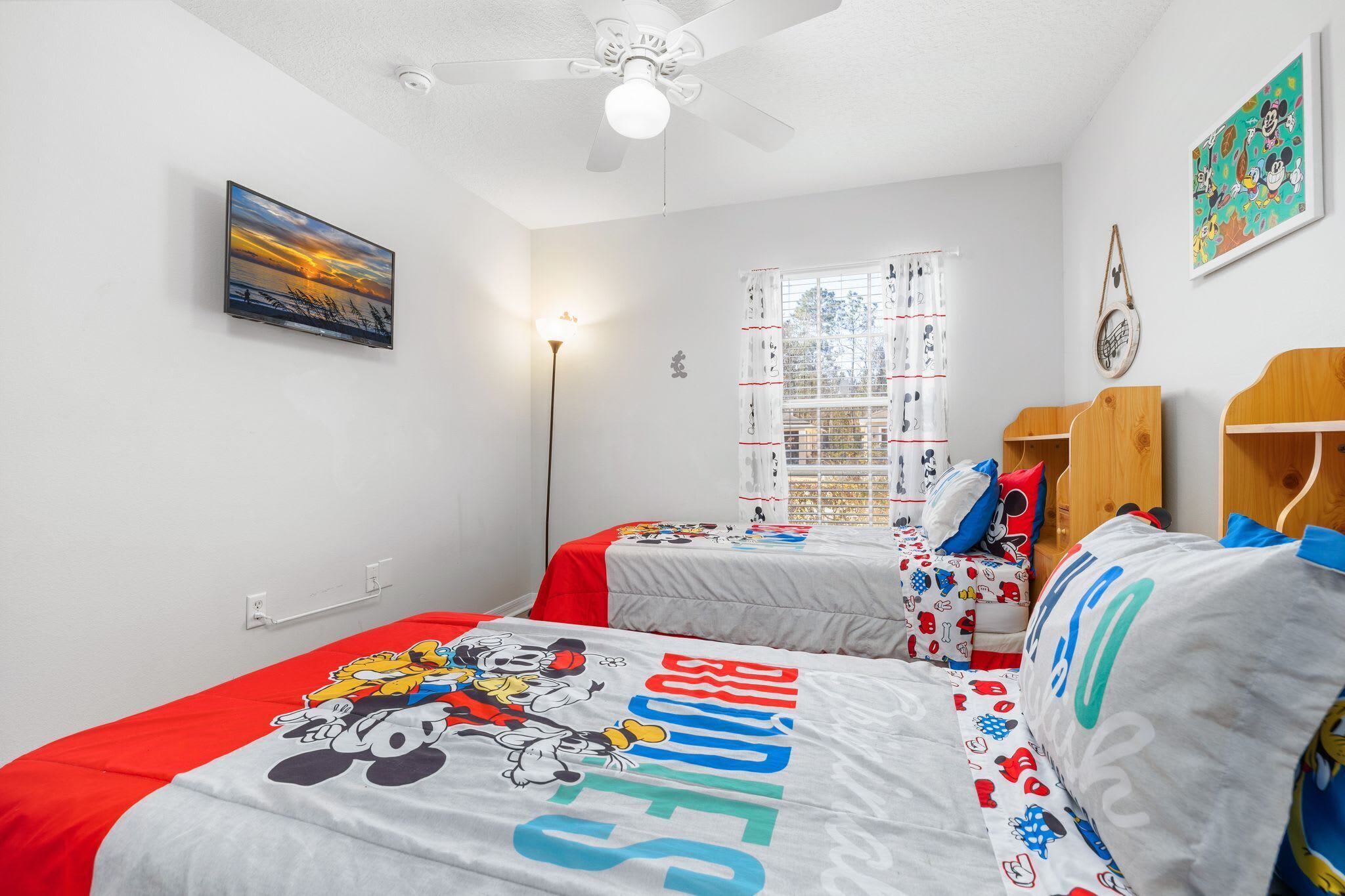 Un dormitorio con dos camas, ventilador de techo, televisor, ventana con cortinas y un cuadro en la pared.