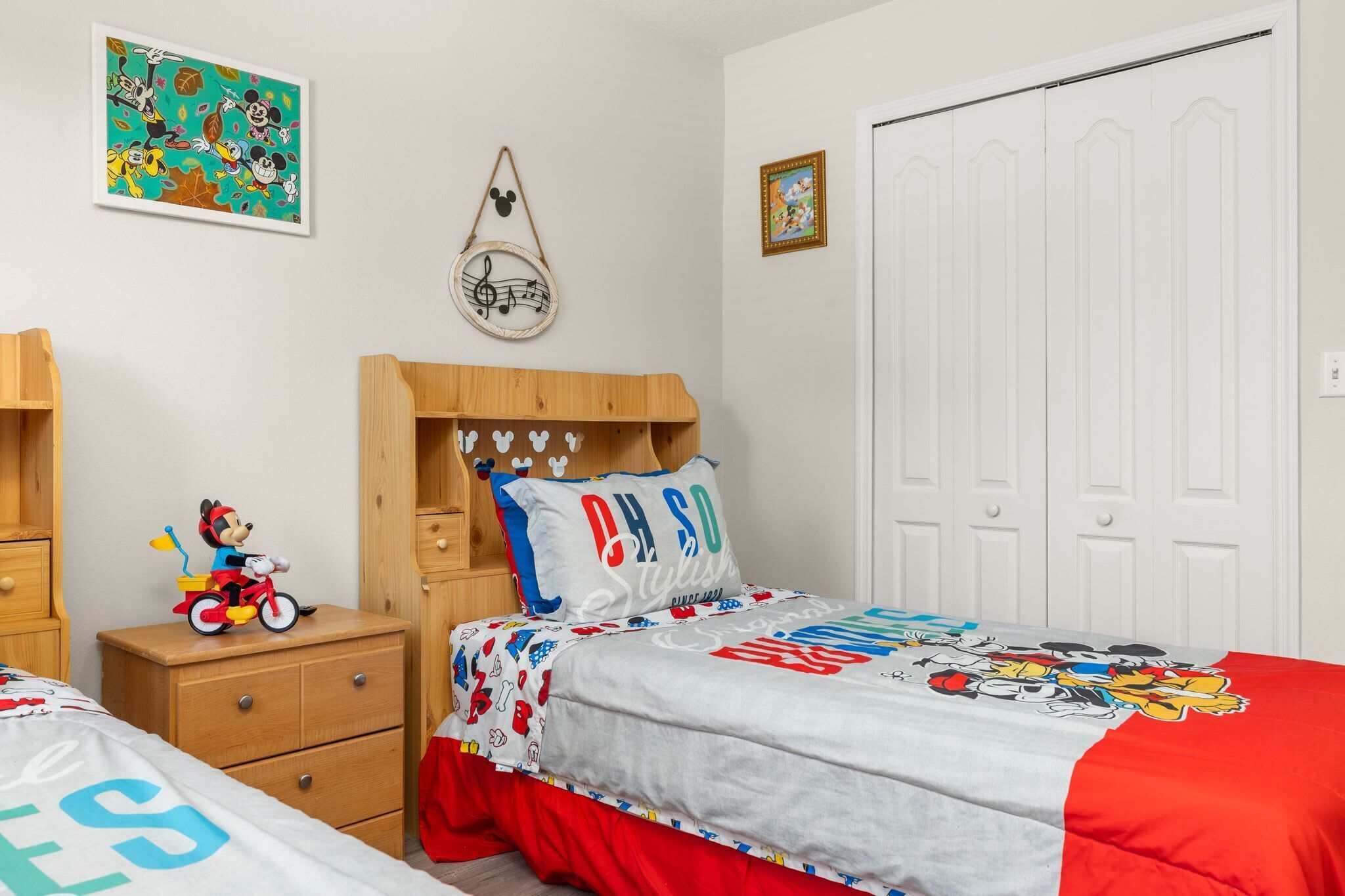 A child's bedroom with a bed, nightstand, dresser, and wall decorations including a Mickey Mouse sign and a framed picture.