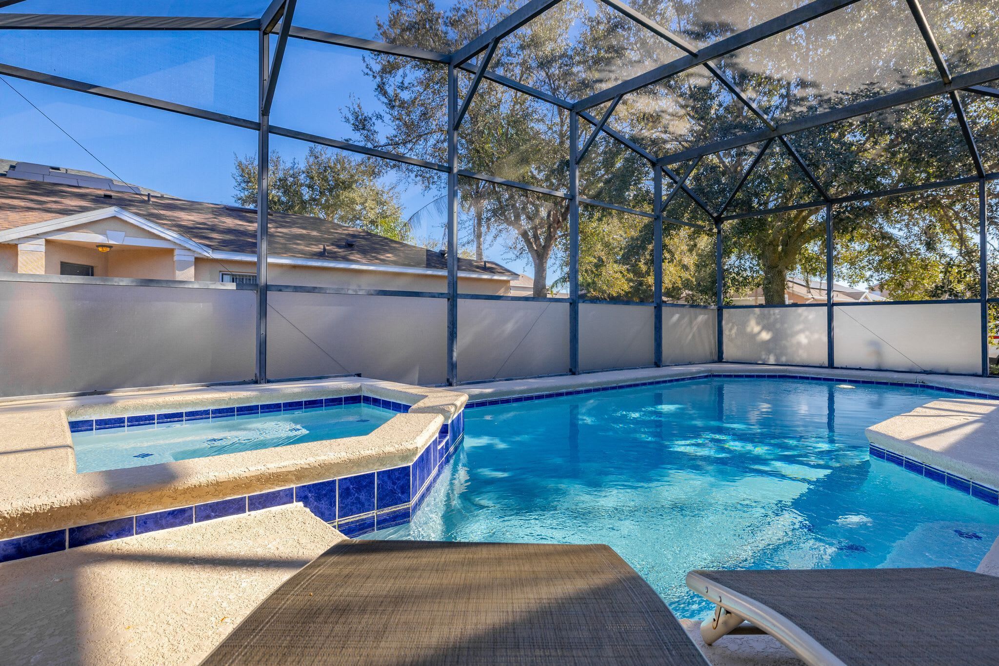 An indoor swimming pool with clear blue water, a hot tub, and a covered area with a view of trees and buildings.