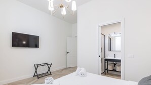 Moderni hotellihuone, jossa on sänky, televisio, kattokruunu ja kylpyhuone, joka näkyy avoimesta ovesta.