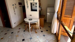 Een kleine keuken met een koelkast, een magnetron en een spoelbak. Er staat een eettafel met twee stoelen en een klein spiegel aan de muur.