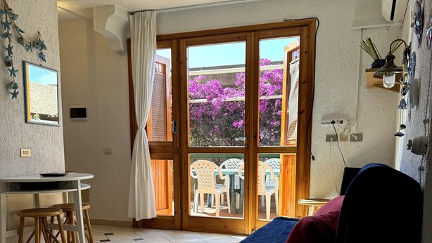 Een kamer met een bed, een bureau en een stoel. Er is een raam met uitzicht op paarse bloemen.