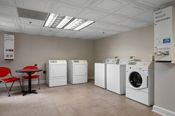 A laundry facility with four washers and dryers, a table with chairs, and a sign listing available detergents. - New Orleans | Urban Retreat | Pool + Hot Tub (New Orleans)