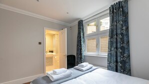 A bedroom with a bed, a window with curtains, a door leading to a bathroom, and a wooden floor. - Beautiful apartment in the heart of Mayfair (London)