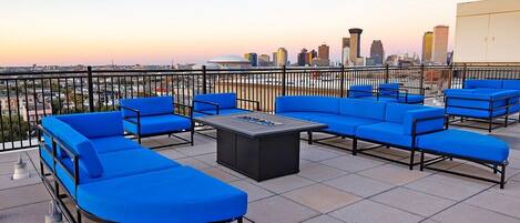 A rooftop terrace with blue seating, a fire pit, and a city skyline view.