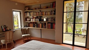 Een kamer met een groot bed, een stoel, een bureau met een lamp en een boekenplank vol boeken. Er is een groot raam met uitzicht op bomen.