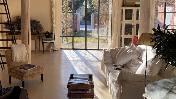 Een ruime kamer met houten balken, een groot raam, een witte bank, een kleine tafel en een stoel.