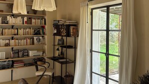 Een knus interieur met een houten plafond, een boekenplank vol boeken, een bureau met stoel en een groot raam met gordijnen.