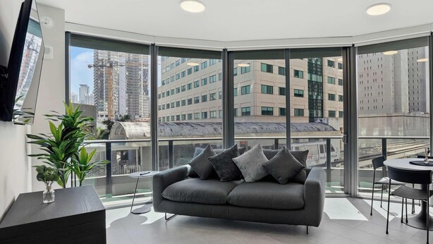 Ein modernes Wohnzimmer mit einem grauen Sofa, einem kleinen Beistelltisch, einer Topfpflanze und einem großen Fenster mit Blick auf die Stadt.