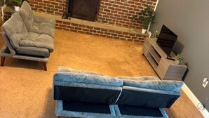 A living room with a brick fireplace, a grey sofa, a striped sofa, and a TV on a wooden stand.