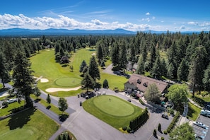 An aerial view of a golf course with a clubhouse, surrounded by dense forest and mountains in the background.