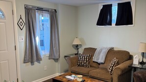 Una sala de estar con un sofá marrón, una mesa de centro de madera, una lámpara y una ventana con cortinas.