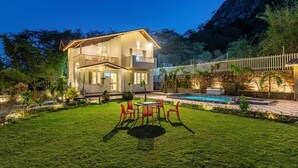 A two-story house with a swimming pool, surrounded by a well-maintained lawn and a stone wall. - Alaya Stays Ramgarh Colonial 2BHK Villa (Jamwa Ramgarh)
