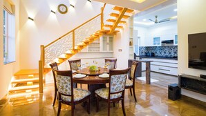 A dining area with a wooden staircase, a round table with chairs, and a kitchen in the background. - Alaya Stays Ramgarh Colonial 2BHK Villa (Jamwa Ramgarh)
