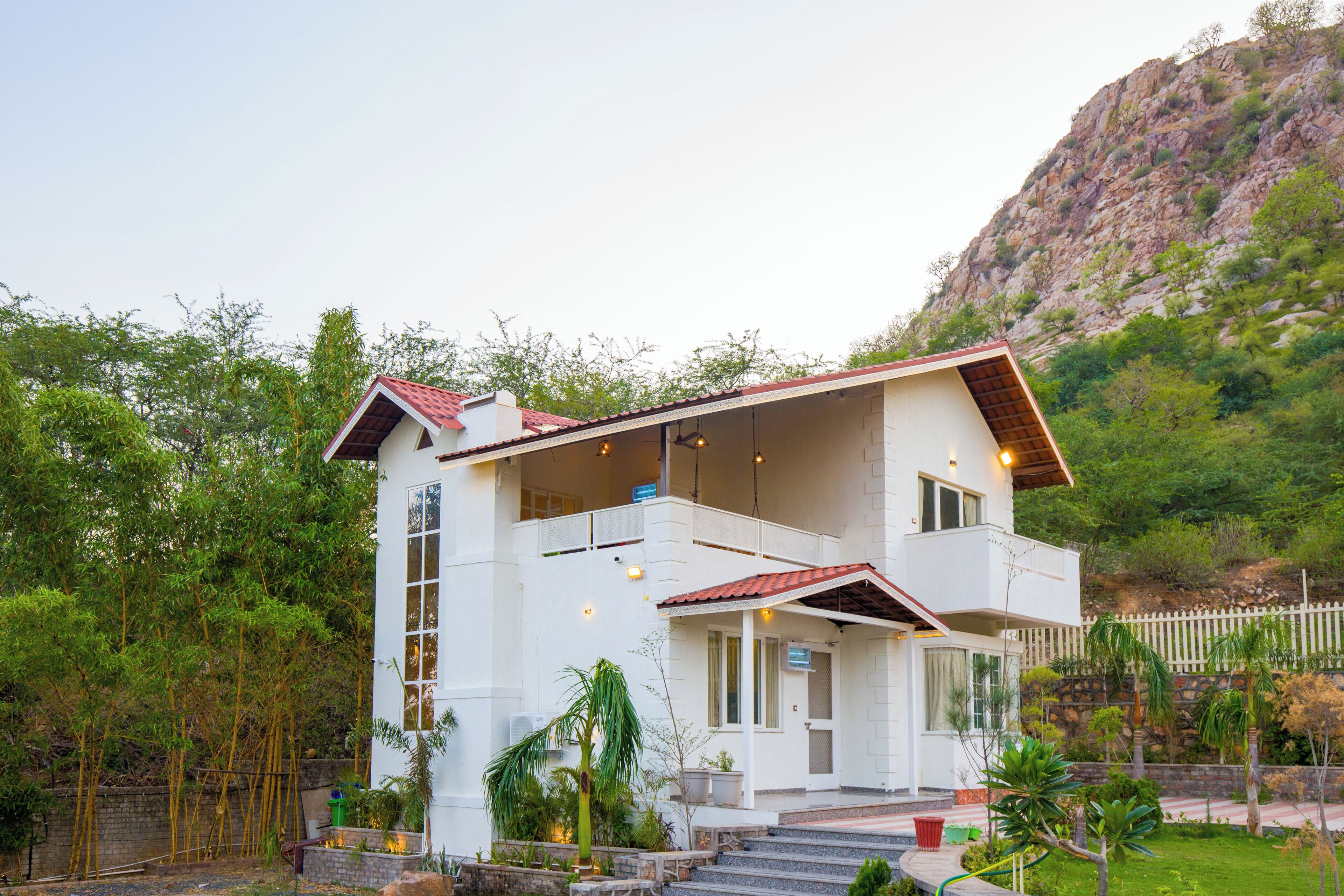 Una casa de dos pisos con balcón, rodeada de vegetación y una ladera.