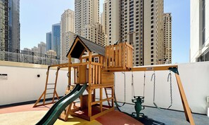 A children's play structure with a slide and swings, set against a backdrop of high-rise buildings. - LUX Contemporary Suite Marina View 5 (Dubai)