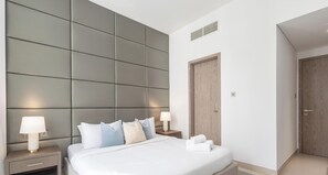 A modern bedroom with a large bed, padded headboard, bedside lamps, a wooden door, and a small nightstand. - LUX Contemporary Suite Marina View 5 (Dubai)