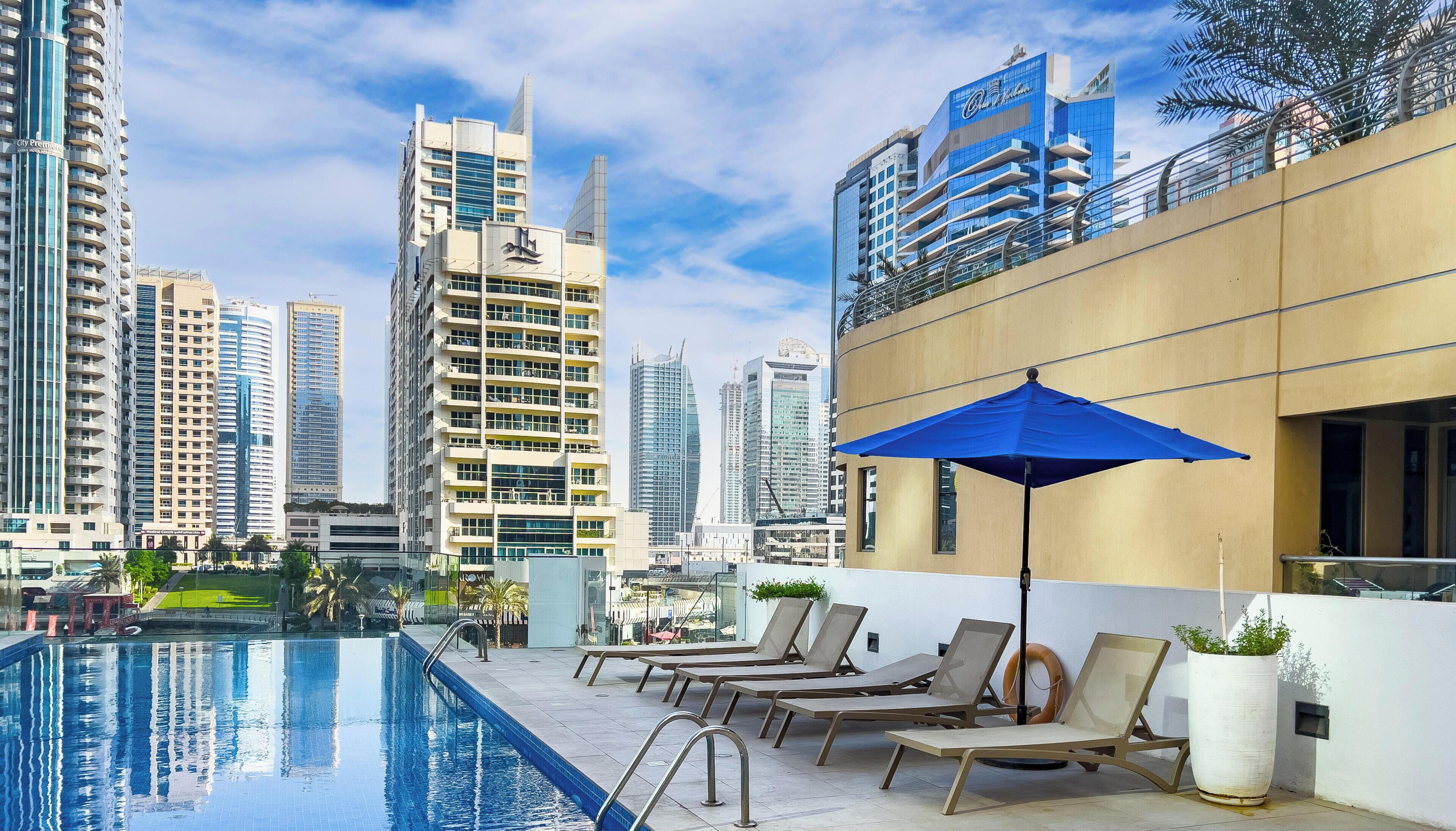 Un área de piscina en la azotea con sillones de descanso y una sombrilla azul, con vistas a un paisaje urbano de edificios altos.