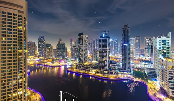 A nighttime cityscape with high-rise buildings, a waterfront promenade, and a river reflecting lights. - LUX Contemporary Suite Marina View 5 (Dubai)